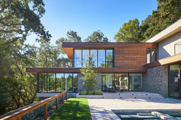 Exterior view of a contemporary multi-level home set among mature trees, with extensive floor-to-ceiling glazing, warm horizontal timber cladding, dark stone wall sections, and broad paved terraces surrounding a partially visible pool. The projecting upper volume and low-slung rooflines create a strong modern composition integrated with the wooded site.