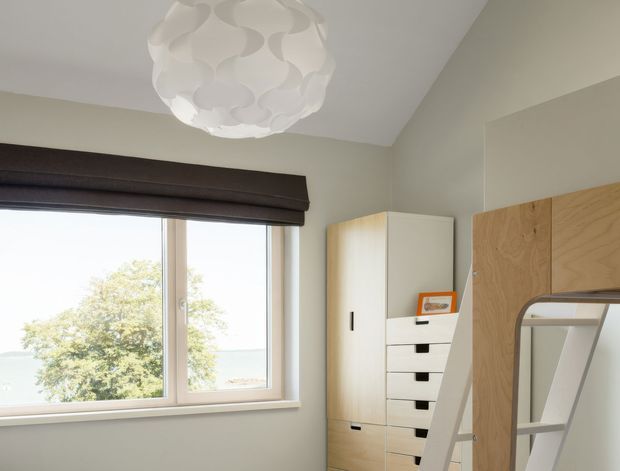 Bright, compact bedroom interior with a large window, dark Roman blind, pale neutral walls, light timber built-in storage, and a partially visible loft-style bed or ladder. A sculptural white pendant light hangs from the ceiling, and leafy outdoor scenery is visible through the window.