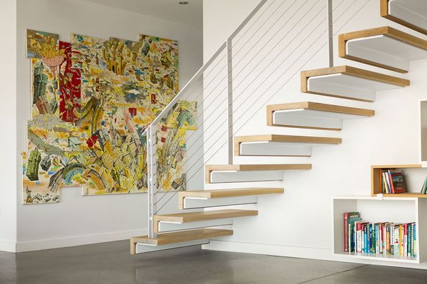 A bright contemporary stairway with floating timber treads and white stringers, supported by a slim metal cable railing. The staircase rises beside a white horizontally panelled wall with integrated open shelving filled with books. A large colourful abstract artwork occupies the adjacent wall, while the room has a smooth grey concrete floor and a clean minimalist fit-out.