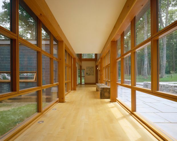 A long, bright glazed corridor or gallery with extensive floor-to-ceiling timber-framed windows on both sides, warm honey-coloured timber flooring and structural posts, and a smooth pale ceiling. The corridor connects to another interior space at the far end and overlooks a landscaped wooded setting with a stone-paved terrace outside. The design emphasizes transparency, natural light, and strong indoor-outdoor connection.