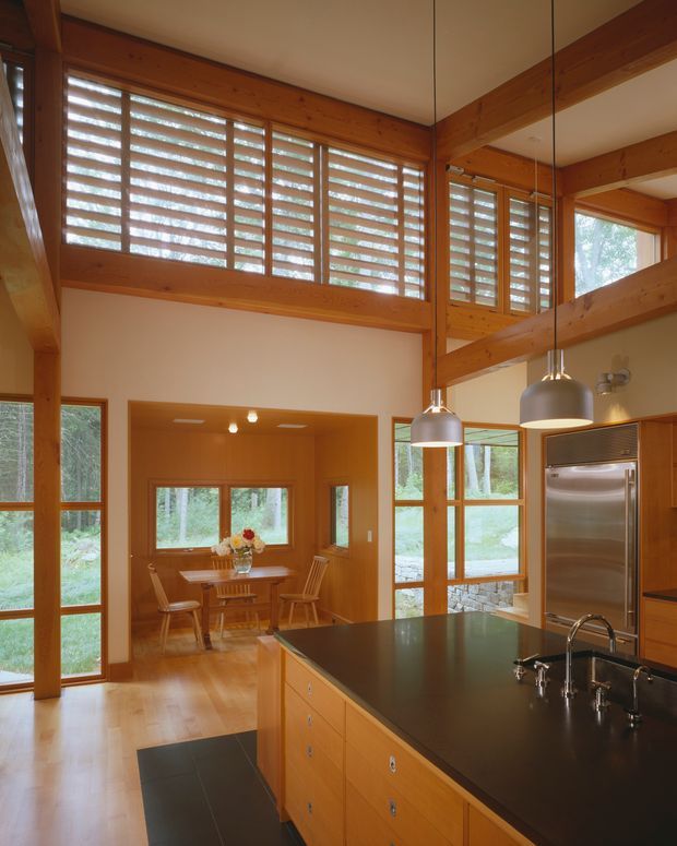 A warm, double-height kitchen and dining area with extensive timber framing and joinery. The kitchen features a large dark benchtop island with an integrated sink and stainless-steel appliances, while a compact dining nook sits beyond. Clerestory windows with horizontal timber louvres bring in filtered natural light, and a large glazed opening provides views to greenery outside. Pendant lights hang above the island.