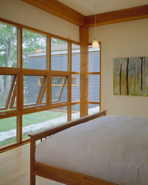 Bright bedroom with a simple timber bed positioned beside extensive floor-to-ceiling glazing. The room features exposed timber structural beams and posts, timber-framed awning windows, pale walls, a light timber floor, a minimal pendant light, and an abstract wall artwork. A landscaped courtyard and adjacent shingle-clad building are visible outside.