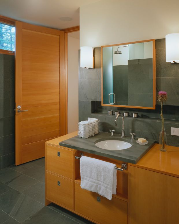 A warm, well-appointed bathroom with a floating timber vanity, integrated drawers and side cabinetry, a grey-green stone benchtop, undermount white basin, wall-mounted chrome tapware, and a large timber-framed mirror. Dark grey-green tiled walls and floor contrast with the honey-toned timber door and joinery. Wall sconces flank the mirror, while a towel rail, toiletries, and a small vase add functional and decorative details.