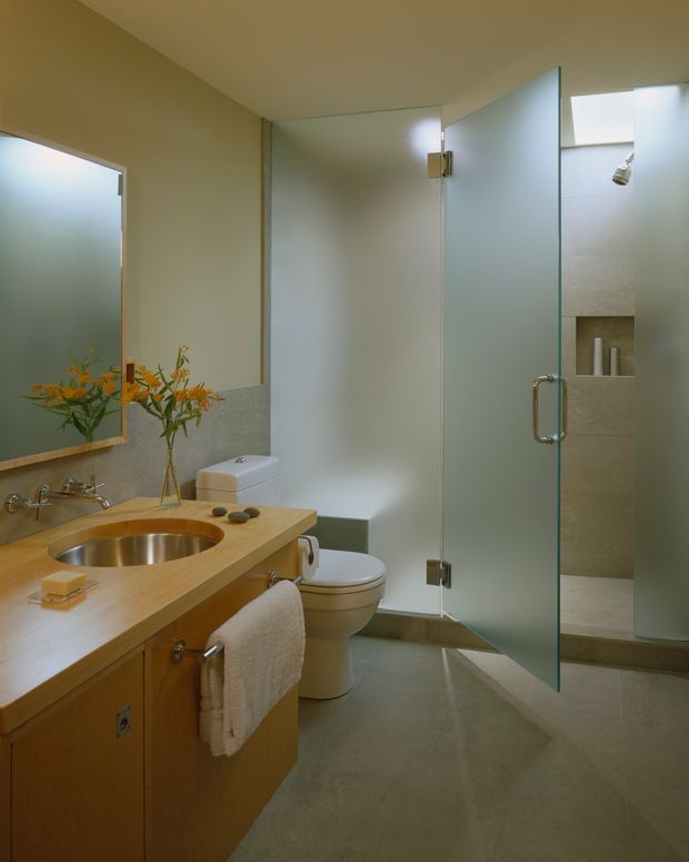 A warm, modern bathroom with a timber vanity, integrated round metal sink, wall-mounted mirror, toilet, and a partially frosted glass shower enclosure. The shower has a recessed niche with small cylindrical containers, and the room features pale stone or concrete-look finishes with brushed metal hardware and soft natural light.