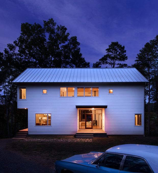 A two-storey rectangular house photographed at dusk, with a pale horizontal weatherboard facade, symmetrical arrangement of windows, a central glazed entry beneath a small projecting awning, and a broad standing-seam metal gable roof. Warm interior lighting glows through the windows and entry doors, contrasting with the deep blue evening sky and surrounding trees. A parked car occupies the lower foreground.