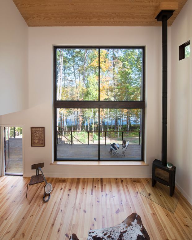 A bright, double-height living space with a very large timber-framed picture window overlooking a wooded landscape and deck. The room has pale timber plank flooring, a warm timber-clad ceiling, light plaster walls, a black freestanding wood-burning stove, and sparse rustic furnishings including a cowhide rug and lounge chairs visible outside.