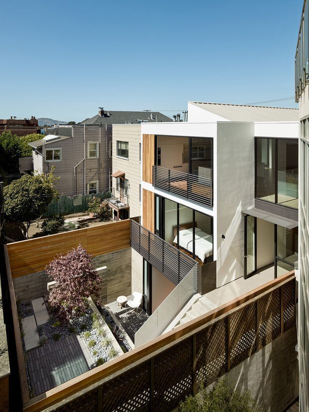 Elevated exterior view of a contemporary multi-level residential building in a dense urban neighbourhood. The white rendered facade features large glazed openings, warm vertical timber accents, projecting balconies with dark metal balustrades, and a flat-to-low-pitched roof form. A landscaped courtyard and timber privacy fencing are visible below, with neighbouring homes in the background.