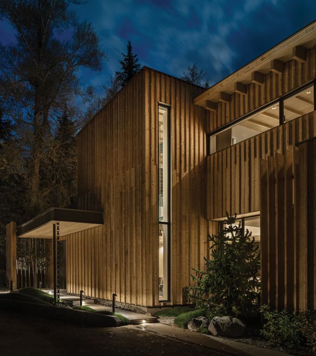 Dusk exterior view of a contemporary multi-level residence set among mature trees. The building features extensive vertical timber cladding, projecting rooflines, deep eaves, tall narrow glazing, and a covered entry walkway with warm interior lighting visible through the windows. A landscaped path, rocks, shrubs, and small trees surround the dark timber facade.