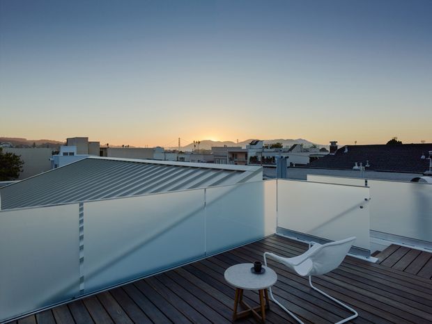A contemporary rooftop terrace at sunset, with dark timber decking, frosted glass privacy screens, white rendered parapet walls, and a minimalist white lounge chair beside a small round table. Low-rise urban rooftops and distant hills are visible beyond the terrace.