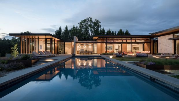 A wide dusk view of a low-slung contemporary residence arranged around a long rectangular swimming pool. The home features flat rooflines, extensive glazing, timber or dark vertical screening, covered outdoor living areas, warm interior lighting, and landscaped courtyards. The pool dominates the foreground and reflects the house and evening sky.