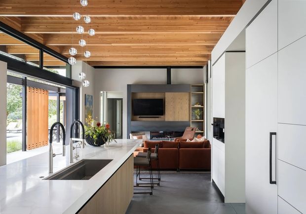 A bright, open-plan contemporary kitchen opening directly into a living area. The kitchen features a long white island with an integrated dark sink, multiple arched black taps, pale wood cabinetry, and tall white built-in storage. The adjoining living room has a rust-coloured sofa, dark media wall, and timber shelving. Black-framed glazing, a warm timber-lined ceiling, polished concrete flooring, and spherical pendant lights create a refined modern interior.