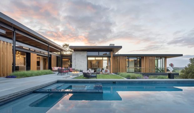 Contemporary single-storey residence arranged around a landscaped courtyard and swimming pool. The home features low-slung flat rooflines, extensive floor-to-ceiling glazing, dark metal-framed windows, warm vertical timber screening, and pale stone or rendered wall surfaces. Outdoor seating and a covered entertaining area face the pool, with an open landscape beyond the property.
