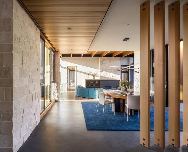 Bright contemporary open-plan interior viewed from an entry or hallway, with a textured masonry feature wall, polished concrete floor, warm timber slatted ceiling, and timber-framed glazing. A dining area with a round table and upholstered chairs sits on a deep blue rug, while a turquoise sofa and low cabinetry define the adjoining living space. Vertical timber pivot or sliding panels partially screen the room, and exposed timber beams add architectural character.