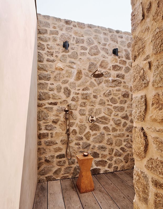 A rustic open shower enclosure with exposed irregular stone walls, timber plank flooring, and aged bronze shower fixtures. A sculptural wooden pedestal with a small basin or accessory stands near the centre. The space has an earthy, warm, natural character and appears open to the sky.