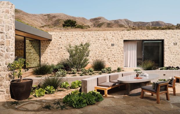 A low-slung stone-clad residence enclosing a landscaped courtyard, with a covered outdoor lounge and dining area in the foreground. The courtyard features built-in concrete or stone seating, a round table, timber-framed armchairs, extensive succulents and drought-tolerant planting, and large dark-framed glazed openings. Dry rocky mountains are visible beyond the wall under a clear blue sky.