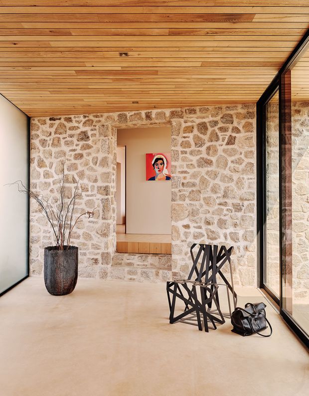A refined interior with exposed irregular stone walls, a warm timber-lined ceiling, pale polished concrete floor, and full-height black-framed glazing along one side. A sculptural black chair and stool, dark ceramic planter with branches, and a small artwork add restrained decorative accents. The view appears to be a passage or gallery-like living space opening toward another room.