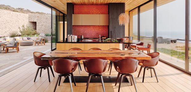 A refined open-plan dining and kitchen pavilion with extensive floor-to-ceiling glazing overlooking a coastal landscape. A long sculptural timber dining table is surrounded by dark leather or wood dining chairs, while the rear kitchen features a dark island, warm timber cabinetry, a red textured splashback, and dark vertical wall cladding. Timber-lined ceilings and pale timber flooring create a warm, contemporary indoor-outdoor atmosphere, with a paved outdoor lounge visible beyond the glazing.