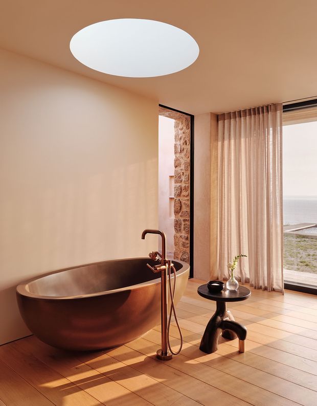 A warm, refined bathroom featuring a freestanding copper-toned oval bathtub, matching floor-mounted bath filler, timber flooring, and a small dark sculptural side table. Full-height sheer curtains frame expansive glazing with a visible coastal horizon, while a stone-clad opening and large circular ceiling light add architectural interest. The space is softly lit with a strong natural, earthy palette.