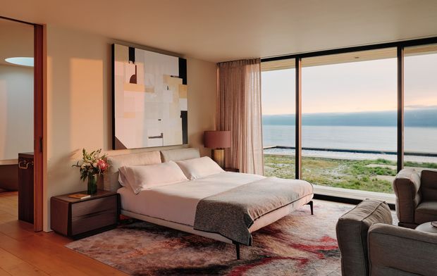 A warm, refined bedroom with a low upholstered bed, dark timber bedside table, abstract wall artwork, layered rug, and floor-to-ceiling glazing overlooking a beach and ocean. A seating area is visible at the right, while a doorway opens to an adjacent room on the left. The room has a contemporary, softly luxurious atmosphere with integrated window treatments and abundant natural light.