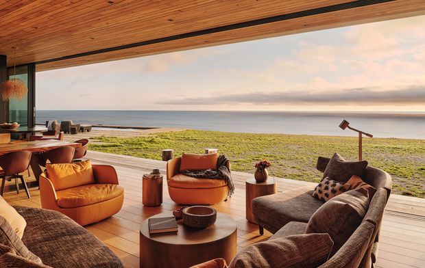 A warm, open-plan coastal living room opening directly onto a timber deck with expansive ocean views. The room features a timber-lined ceiling with recessed linear detailing, full-height glazing, timber flooring and deck surfaces, a dining area, and richly upholstered tan and brown lounge furniture arranged around a round timber coffee table. A beach and calm water are visible beyond the outdoor opening.