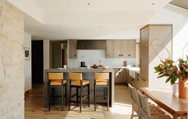 Warm, light-filled open-plan kitchen and dining area with a central island and three timber bar stools. The kitchen features pale timber cabinetry, a muted stone or concrete-look splashback and benchtops, dark timber flooring, recessed ceiling lighting, and a substantial light-stone masonry feature wall. A dining table and woven dining chairs sit beside large windows, with indoor greenery adding a natural accent.