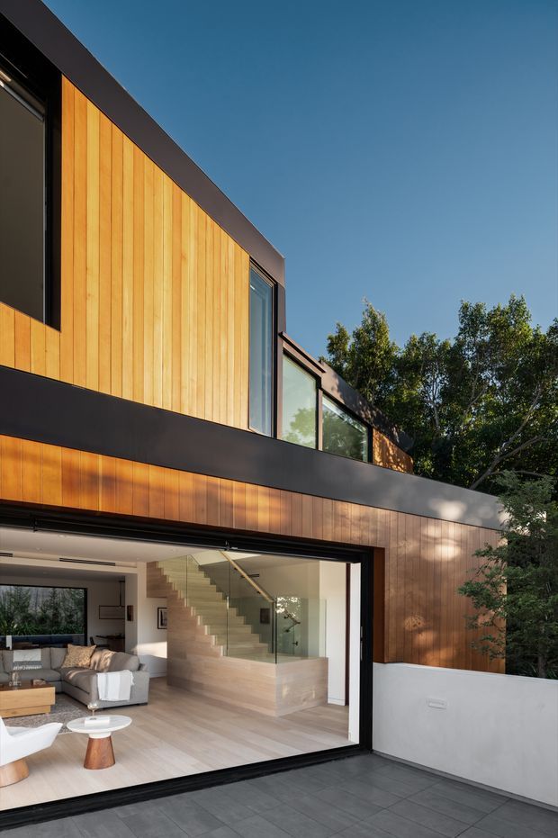 Exterior view of a contemporary multi-level home with warm vertical timber cladding, dark metal framing and trim, large glazed openings, and a partially visible open-plan living room with a floating staircase. A paved terrace occupies the foreground, with mature trees beside the building.
