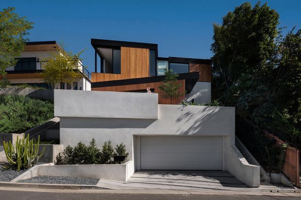 Street-facing view of a contemporary multi-level residential house on a sloping site. The home features strong geometric forms, flat rooflines, dark window frames, warm vertical timber cladding, smooth white rendered/concrete walls, a projecting upper volume, balconies, and an integrated double garage. Dense landscaping surrounds the stepped driveway and retaining walls.