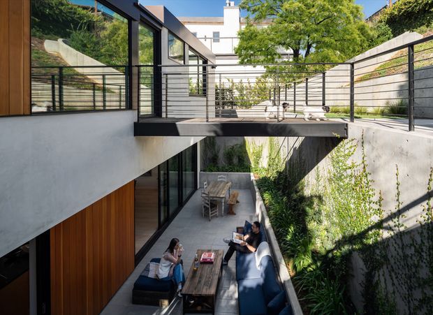 A contemporary multi-level residential courtyard viewed from above, with a sunken outdoor lounge and dining area below a cantilevered deck or bridge. The architecture combines white rendered walls, warm vertical timber cladding, extensive black-framed glazing, and slender black metal balustrades. Concrete retaining walls and lush planted walls line the narrow courtyard, while an upper terrace and neighbouring urban structure are visible beyond.