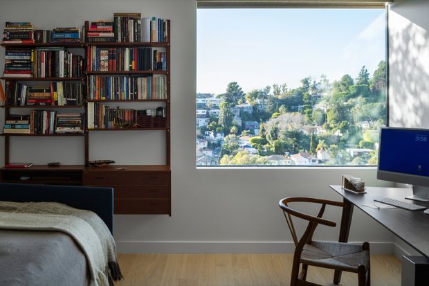 A compact, bright bedroom with a built-in wall of dark timber bookshelves and drawers, a partially visible bed, and a dedicated desk positioned beside a large picture window. The window overlooks a leafy residential hillside, while light timber flooring and neutral walls create a calm, contemporary workspace-and-bedroom setting.