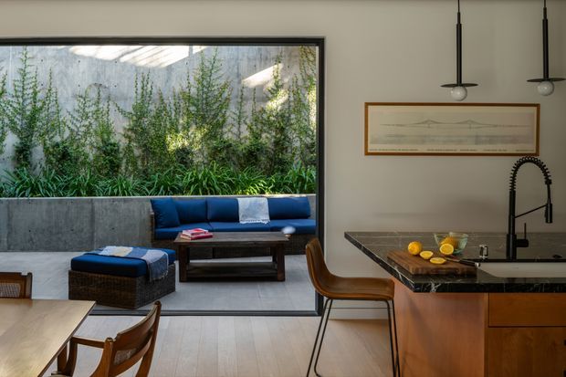 A refined open-plan kitchen and dining area beside a sunken living space. The room features a dark stone kitchen island with warm timber cabinetry, a black gooseneck tap, pendant lights, timber dining furniture, and a large black-framed window overlooking a planted concrete courtyard. The living area includes a deep blue built-in sofa and ottoman, creating a warm contemporary interior with strong indoor-outdoor connection.