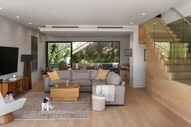 A contemporary open-plan living room with a large grey sectional sofa, timber coffee table and media console, patterned area rug, and floor-to-ceiling glazing overlooking greenery. A timber-clad staircase with clear glass balustrade forms a prominent architectural feature on the right. The room has recessed ceiling lighting, neutral painted walls, warm timber flooring, and a bright, relaxed atmosphere.