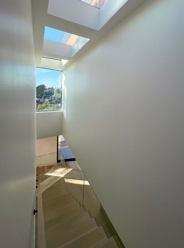 A bright modern stairwell viewed from above, featuring a minimalist white plastered enclosure, pale timber stair treads, clear glass balustrades, and multiple overhead skylights. A large window provides views toward a leafy residential landscape, while strong sunlight creates geometric shadows across the stairs and landing.