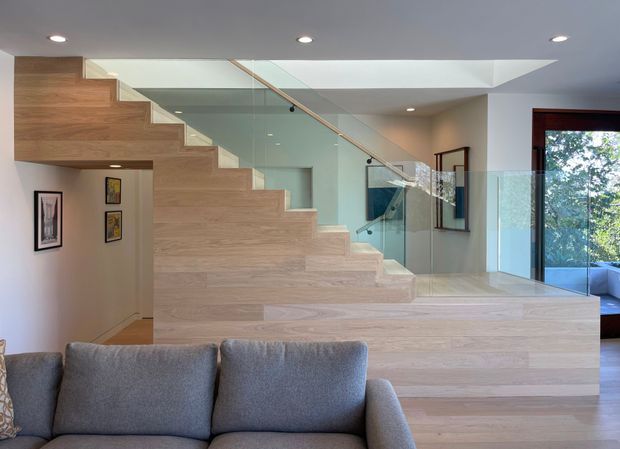 Contemporary open-plan interior featuring a sculptural pale timber staircase with broad integrated treads and a glass balustrade. The stair rises beside a grey upholstered sofa and leads toward a bright landing, with recessed ceiling lights, neutral walls, framed artwork, and large glazed openings overlooking greenery.