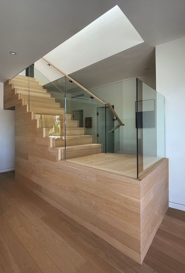 Interior view of a contemporary stairway with broad light timber-clad stair structure and treads, clear glass balustrades, slim timber handrails, and a large rectangular skylight above. The stair rises through a compact double-height or vertically open area, with white walls and timber flooring creating a warm, minimalist composition.