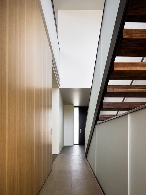 A narrow, minimalist interior hallway with full-height light timber built-in cabinetry along the left wall, smooth white walls and ceiling, and a dark tiled floor. A black-framed door with a tall vertical glazed panel anchors the far end. On the right, a balustrade overlooks or borders a lower level, with exposed dark timber ceiling beams visible above, creating a strong linear architectural composition.