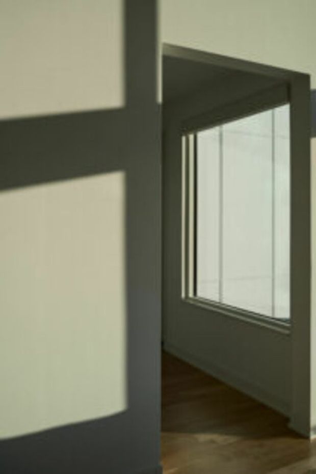 Minimal interior architectural view of a narrow passage or adjoining room with strong geometric sunlight and shadow patterns, pale plaster walls, dark window and door framing, and warm timber flooring. The large glazed opening and crisp rectangular forms create a quiet, contemporary atmosphere.