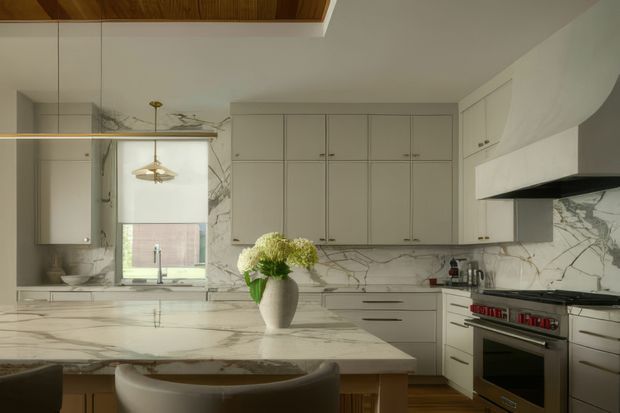A refined contemporary kitchen with extensive pale veined stone surfaces, warm grey shaker-style cabinetry, integrated appliances, a large island with seating, and a timber-lined ceiling detail. A large window and pendant lighting add natural light, while a floral arrangement provides a small decorative accent.