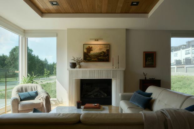 A bright, refined living room with a large cream sectional sofa and armchair arranged around a central fireplace. The fireplace has a white fluted surround and mantel, while the room features a warm timber-panelled recessed ceiling, pale walls, expansive windows, and restrained traditional artwork and furnishings.