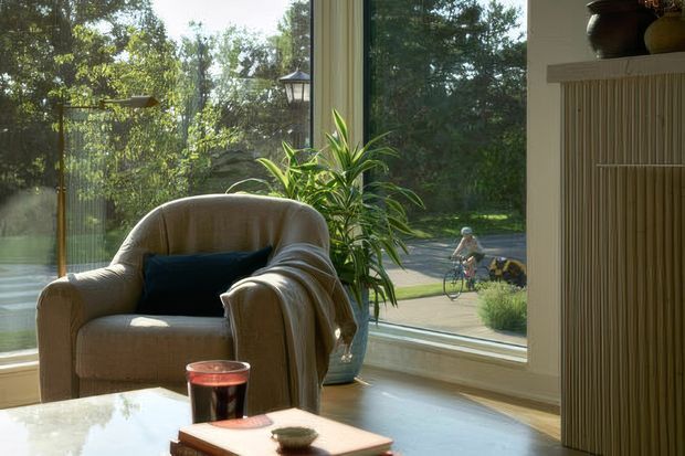 A warmly lit sitting area beside expansive floor-to-ceiling glazing, featuring a plush beige armchair with a dark cushion, a potted indoor plant, and a low coffee table. Vertical timber slatted built-in joinery is visible on the right, while a leafy landscaped exterior can be seen through the windows.