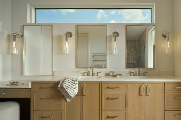 Bright, refined bathroom vanity with a long pale stone benchtop, extensive light timber cabinetry, twin basins, framed mirrors, wall sconces, and a high horizontal window. A separate shower or bathroom area is visible in the right mirror reflection. The room has a clean, warm contemporary-traditional character with abundant natural light.