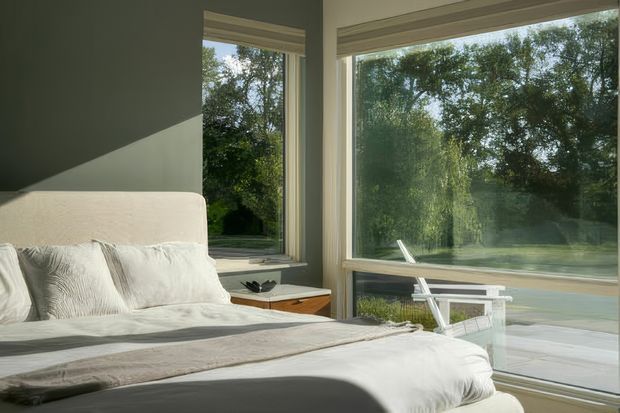 A serene bedroom with a low-profile upholstered bed dressed in white and pale grey bedding. Large corner windows provide abundant natural light and overlook leafy greenery and a terrace or outdoor area. The room has muted grey-green walls, simple roller blinds, a small timber bedside table, and a minimal, contemporary feel.