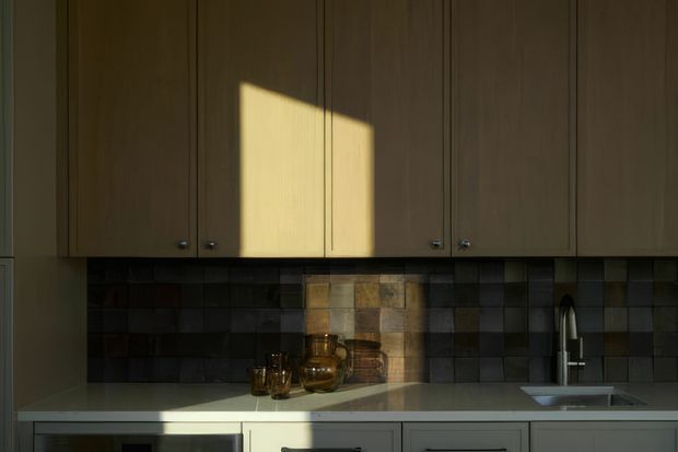 A compact, warmly lit kitchen viewed straight on, with full-height natural timber cabinetry, a pale solid-looking benchtop, dark square tiled splashback, and a stainless steel gooseneck tap over a single-bowl sink. Strong sunlight creates a bright rectangular patch across the cabinetry and benchtop, while simple glassware and a ceramic vessel sit on the counter.