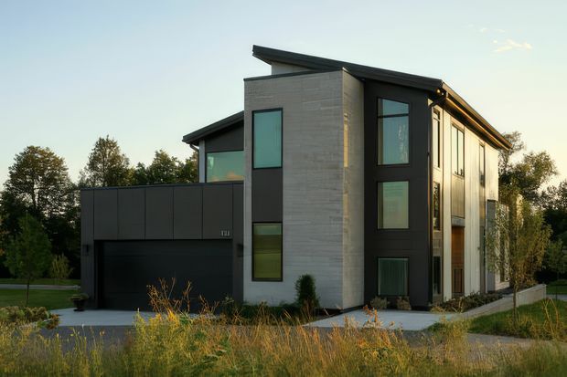 Modern multi-storey residence viewed from the front, featuring a strong geometric composition, a tall central pale masonry volume, dark metal-framed vertical glazing, a projecting flat roof, and a dark attached garage volume. The house sits within a landscaped suburban setting with trees and grasses around a paved driveway.