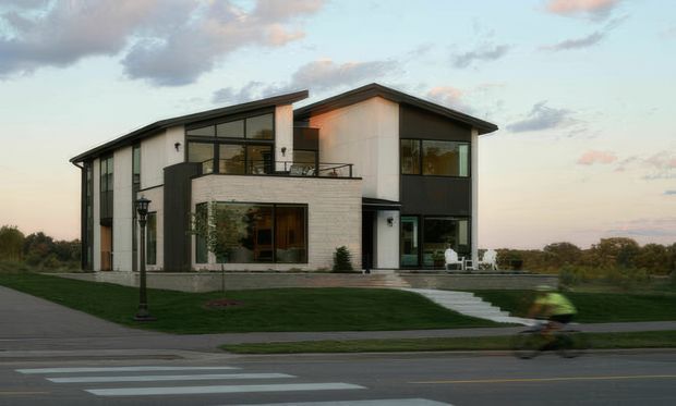 A contemporary two-storey detached residence viewed from across the street at dusk. The home features a strongly articulated gabled roof, contrasting dark and light facade panels, expansive black-framed glazing, a projecting pale masonry-clad ground-floor volume, upper-level balcony, and a broad manicured lawn with a paved path to the entrance. A street and blurred cyclist occupy the foreground.