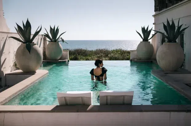 A narrow infinity-edge swimming pool sits between pale rendered building walls and large sculptural planters containing agave plants. Two submerged lounge chairs face the ocean, with a person standing in the turquoise water. The scene has a refined coastal resort character and strong ocean views.