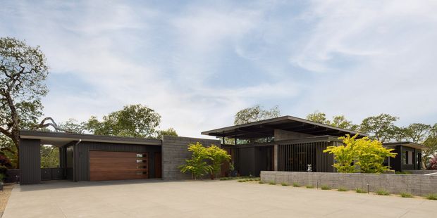 Wide exterior view of a low-profile contemporary residence with dark vertical cladding, broad flat roof planes, exposed concrete block walls, and an integrated double garage with a timber-toned door. The home is approached by a large pale concrete driveway and landscaped with small trees and shrubs.