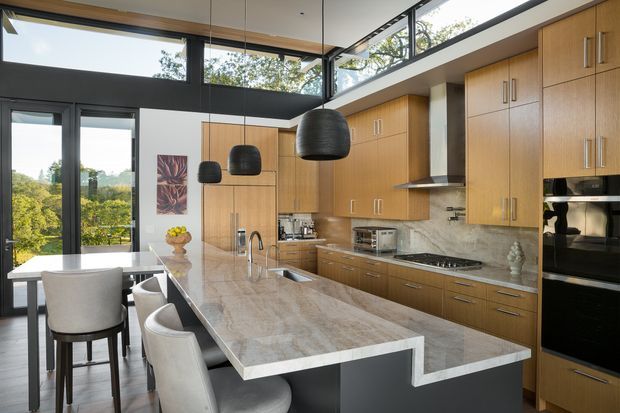 A bright, contemporary kitchen with extensive warm timber cabinetry, a large marble-look island with integrated seating, dark-framed floor-to-ceiling glazing, and black pendant lights. The kitchen opens toward a balcony or terrace with leafy views, while integrated appliances and a stainless-steel rangehood create a refined, functional fit-out.