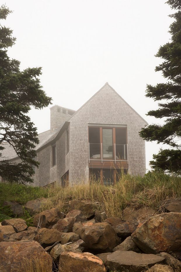 A shingle-clad house viewed from a rocky, grassy slope, framed by mature evergreen trees in misty conditions. The building has a steep gabled roof, a prominent central glazed opening with a narrow balcony and metal railing, and a substantial masonry chimney. The muted natural setting and weathered materials create a rugged coastal or alpine architectural character.