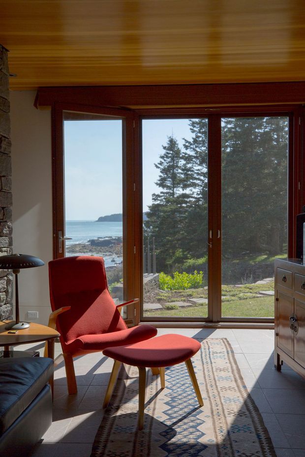 A sunlit coastal sitting area with a red upholstered lounge chair and matching ottoman, positioned beside a small side table and floor lamp. Full-height timber-framed glazing overlooks rocky shoreline, ocean, and trees. The room has a warm timber-paneled ceiling, a stone feature wall at left, tiled flooring, and a patterned runner rug.