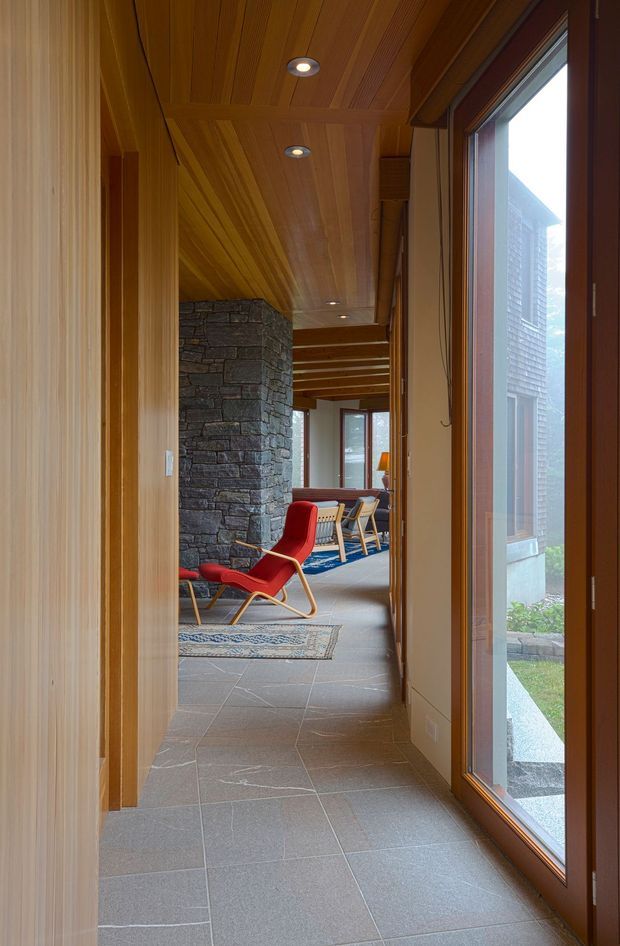 A warm, wood-lined interior hallway leading into a mid-century-inspired living area. The corridor has a richly timber-panelled ceiling and wall trim, large timber-framed glazing on the right, and large grey stone or porcelain floor tiles. Beyond the hallway is a curved dark stacked-stone feature wall, a red lounge chair, a blue rug, and additional seating. The space feels connected to a landscaped exterior through the full-height window.